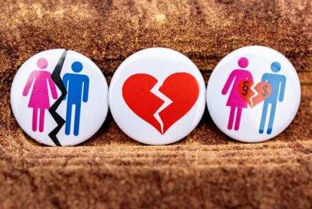 Three pin back buttons show a man and woman couple with a split between them, a broken heart, and a second man and woman couple with a broken heart shown with dollar signs in the heart